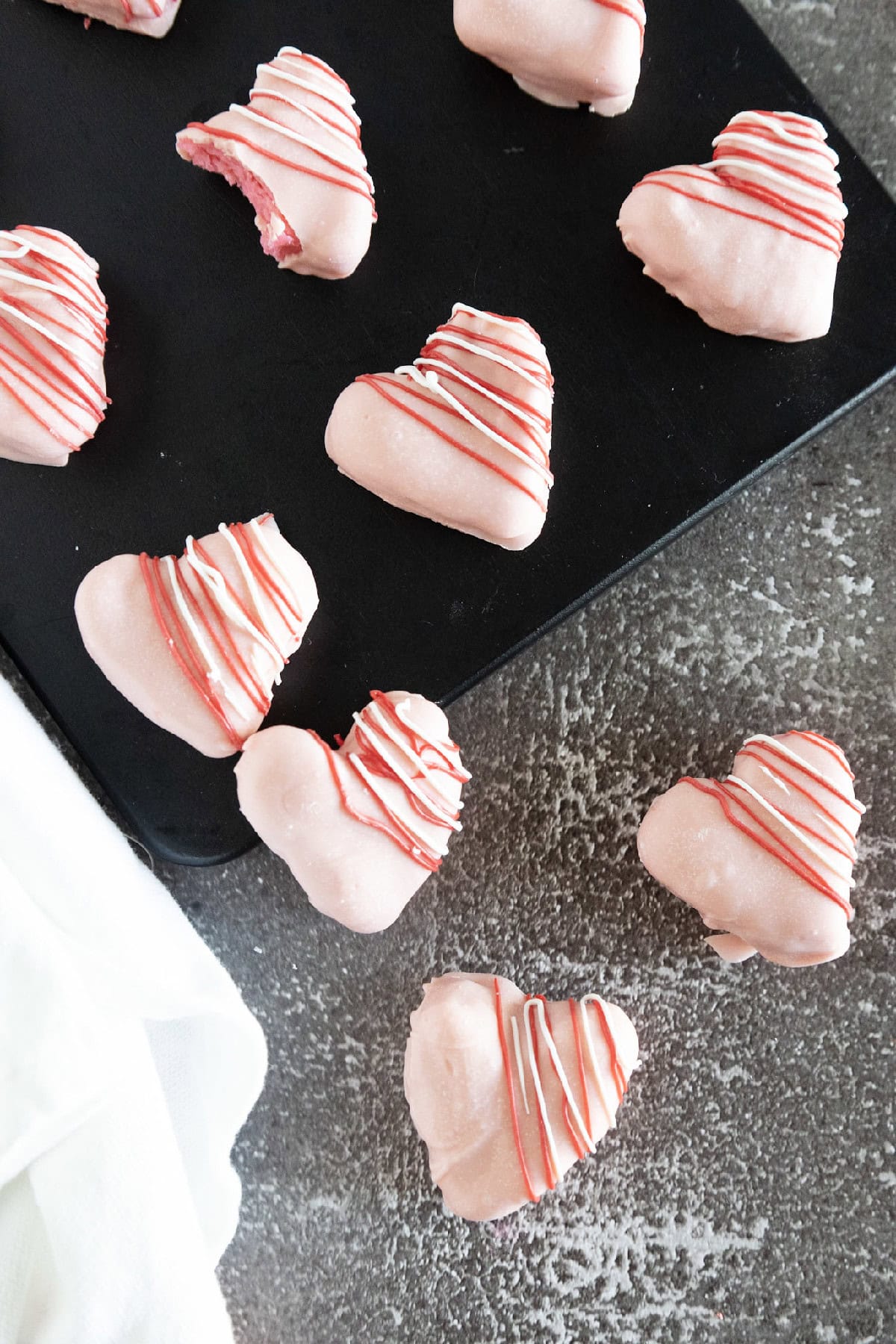 Pink heart-shaped cake truffles coated in candy melts and drizzled with red and white chocolate, arranged on a black serving tray.