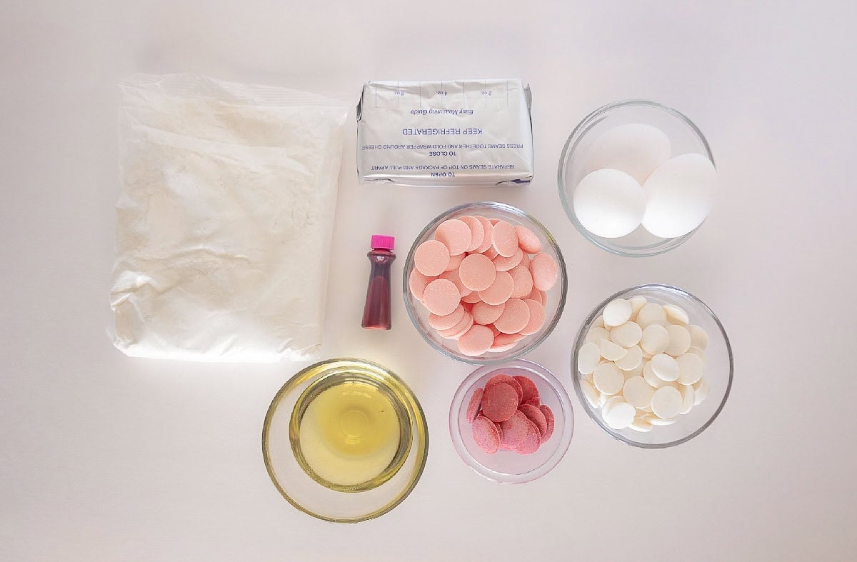 Ingredients for pink cake truffles arranged on a white surface, including cake mix, eggs, cream cheese, oil, pink candy melts, white candy melts, red candy melts, and pink food coloring.
