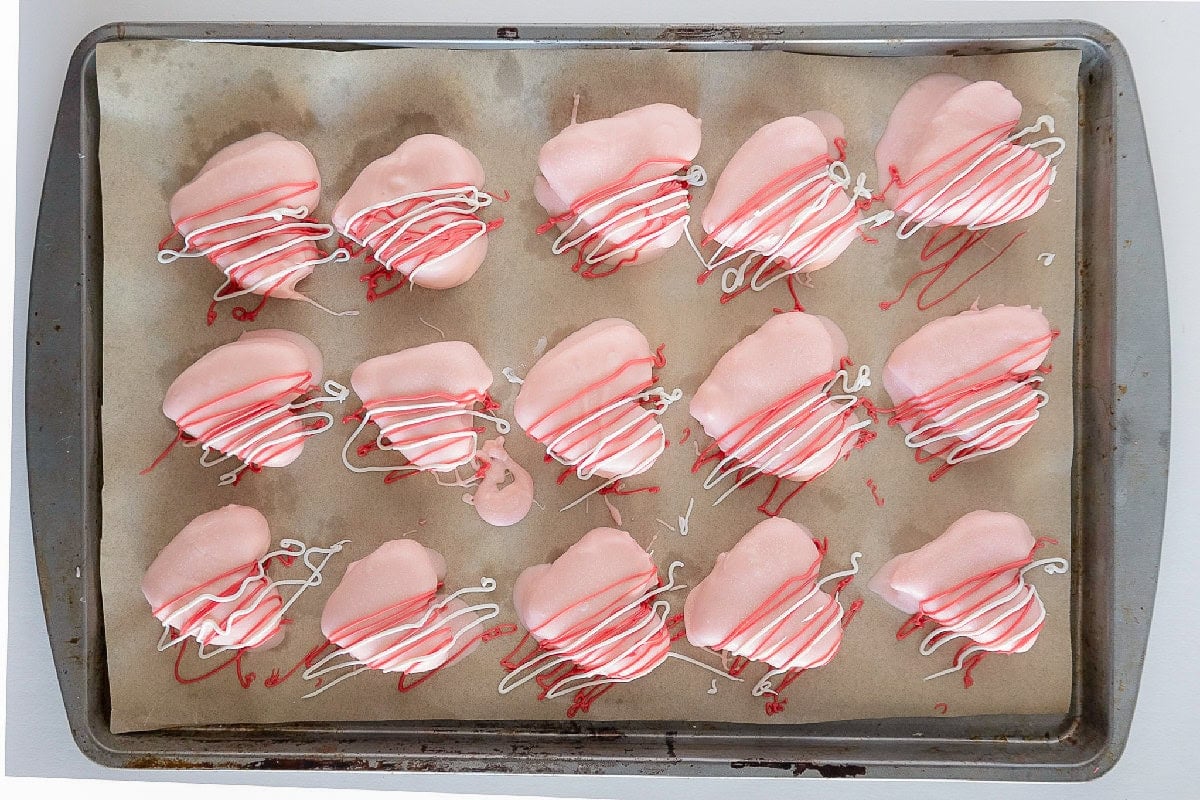 Pink heart-shaped cake truffles decorated with red and white candy drizzle on a baking sheet.