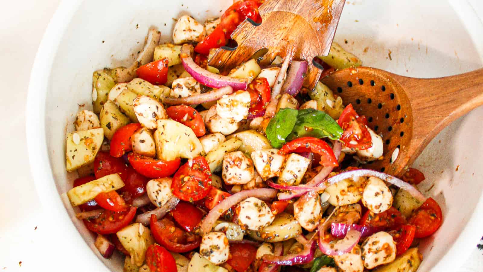 Finished cucumber caprese salad served in a rustic wooden bowl with cucumber chunks, cherry tomatoes, vegan mozzarella pearls, red onion, fresh basil, and rice vinegar dressing.