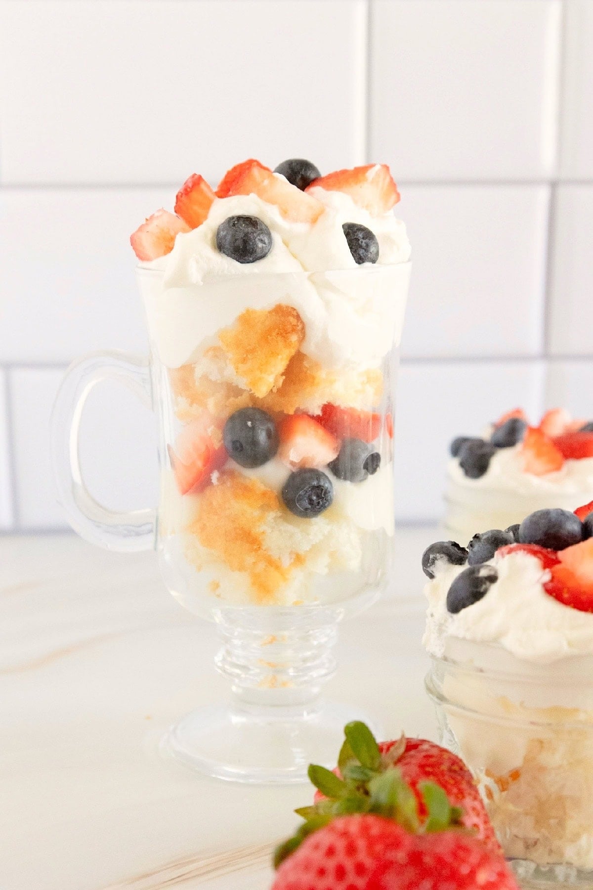 Individual red, white, and blue trifle layered with angel food cake, vanilla pudding, whipped topping, strawberries, and blueberries in a glass mug.