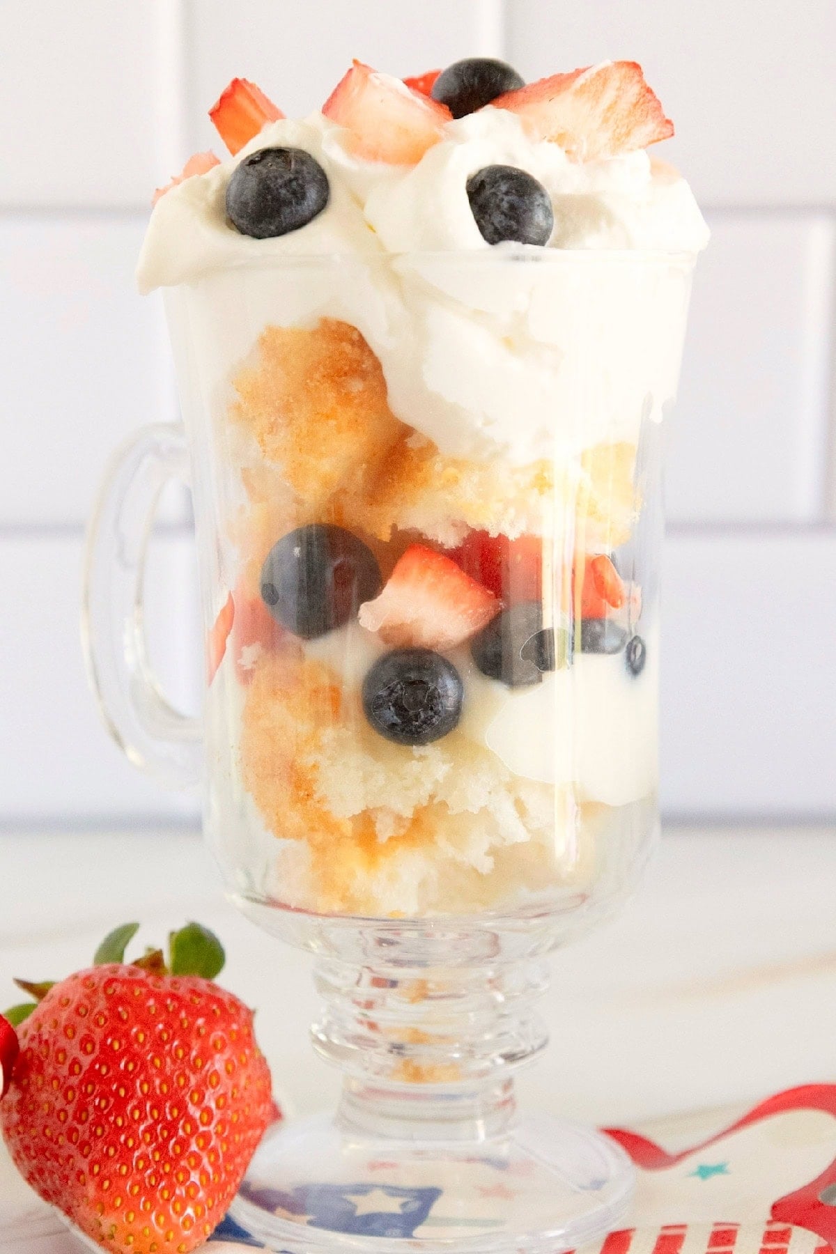 Side view of a red, white, and blue trifle topped with whipped topping, strawberries, and blueberries in a glass mug.