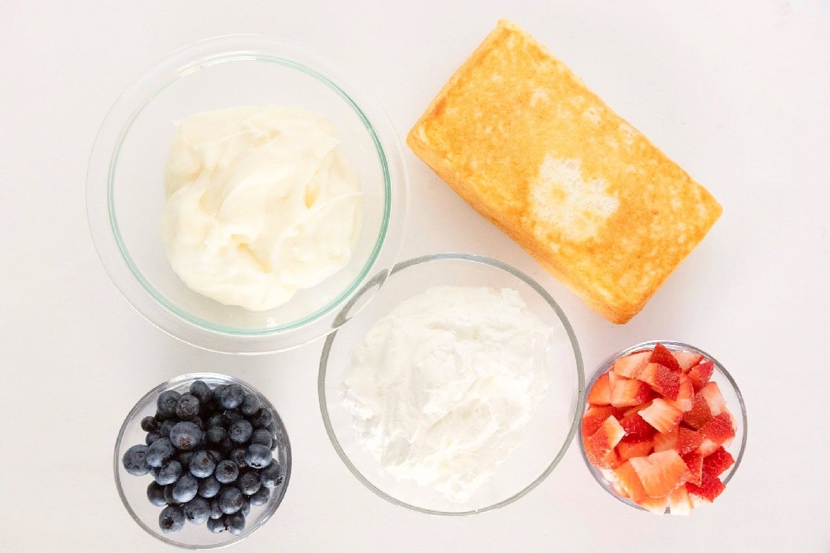 Ingredients for red white and blue trifle including angel food cake, vanilla pudding, whipped topping, fresh blueberries, and chopped strawberries.