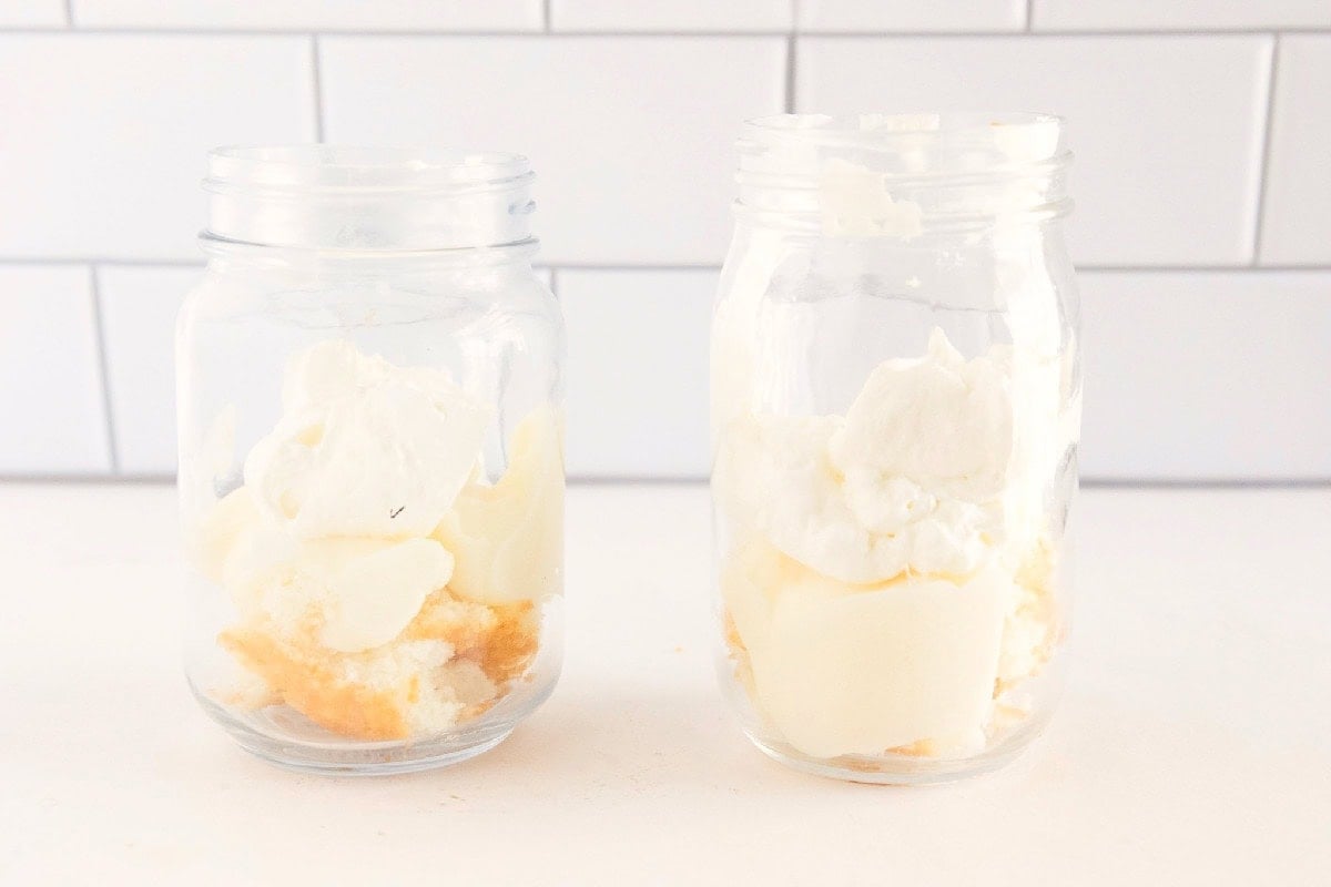 Vanilla pudding and whipped topping added over angel food cake in glass jars for trifle layers.