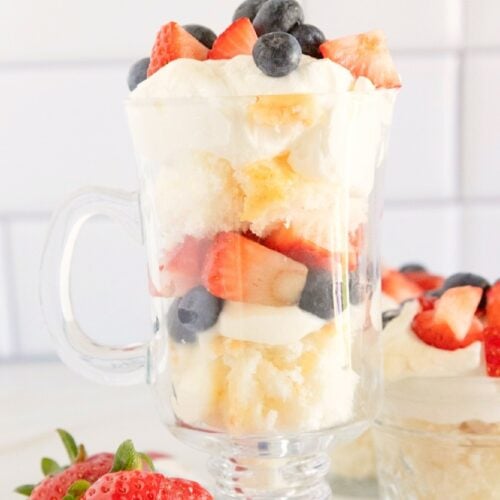 Red white and blue trifle layered with angel food cake, vanilla pudding, whipped topping, fresh strawberries, and blueberries in a glass dessert mug.
