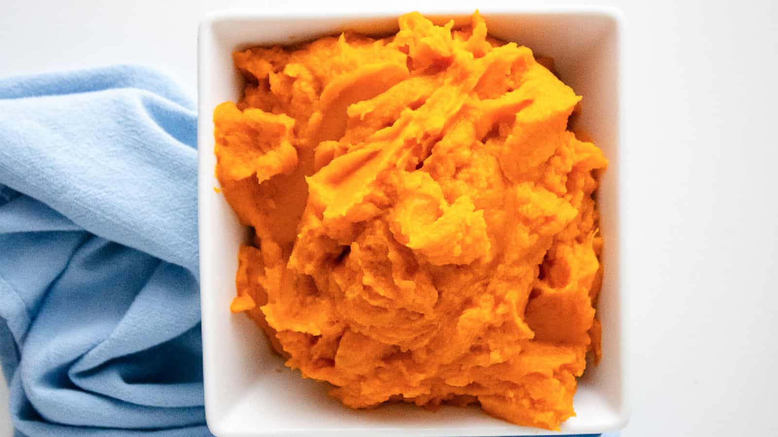 Wide hero image of creamy sweet potato puree in a white bowl with a blue napkin on a white background.
