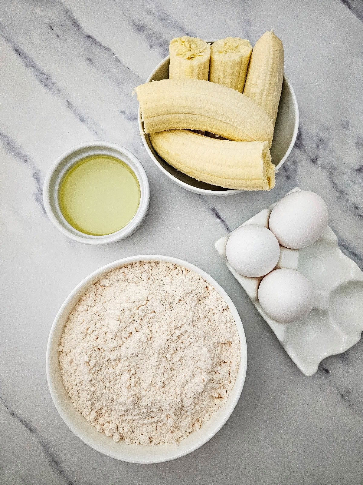 Ingredients for banana bread using yellow cake mix, including yellow cake mix, ripe bananas, eggs, and vegetable oil on a marble countertop.