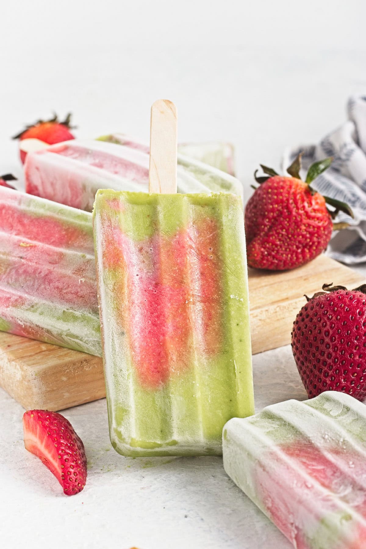Upright matcha popsicle with strawberry layers surrounded by fresh strawberries and frozen popsicles.