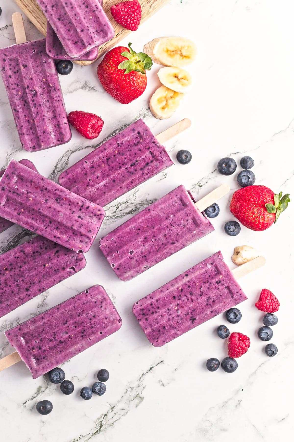 Homemade triple berry smoothie popsicles arranged on a marble surface with fresh strawberries, blueberries, raspberries, and banana slices.