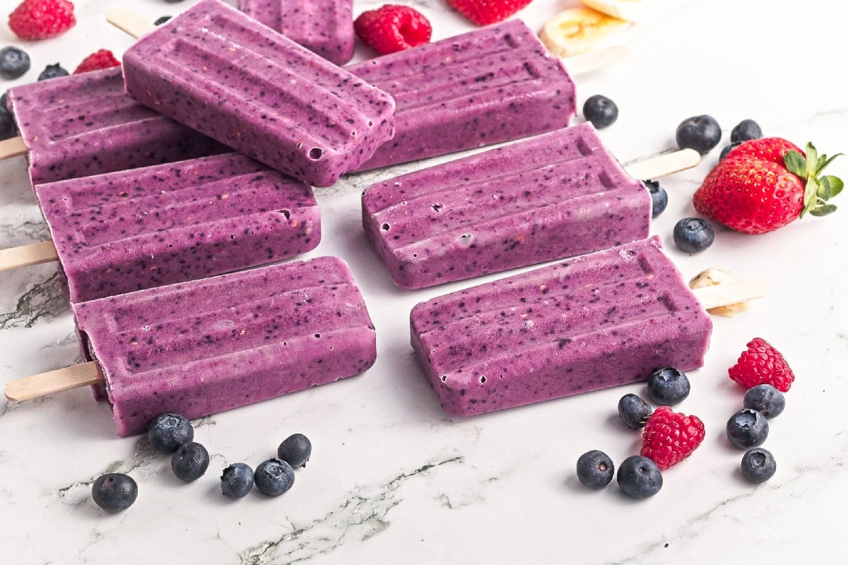 Homemade triple berry smoothie popsicles served with fresh blueberries, raspberries, and strawberries on a marble surface.