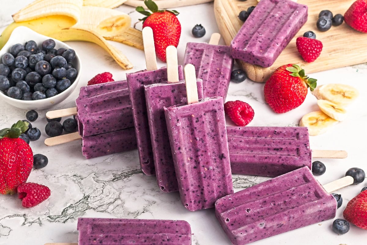 Triple berry smoothie popsicles arranged upright with fresh blueberries, strawberries, raspberries, and banana slices.