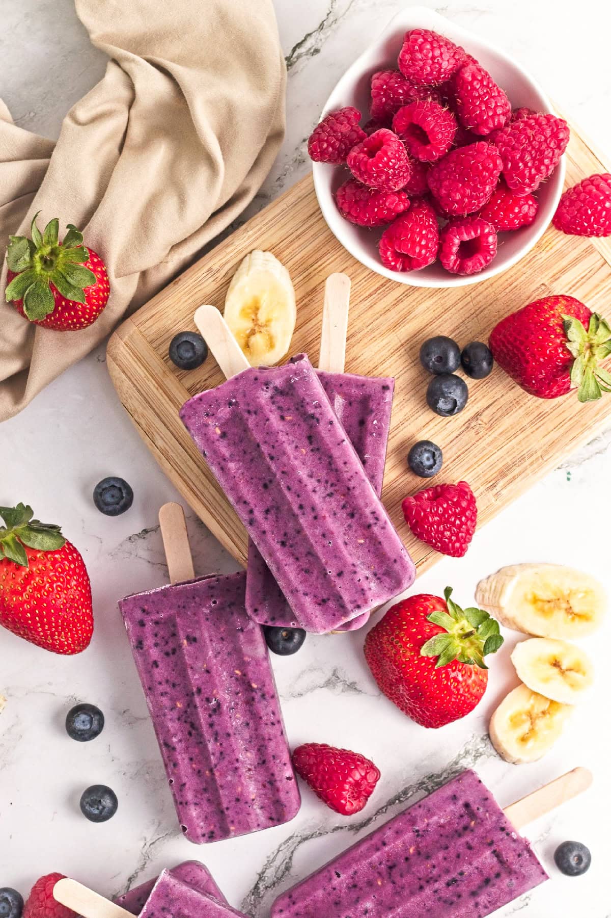 Triple berry smoothie popsicles stacked on a wooden board with fresh strawberries, raspberries, blueberries, and banana slices.