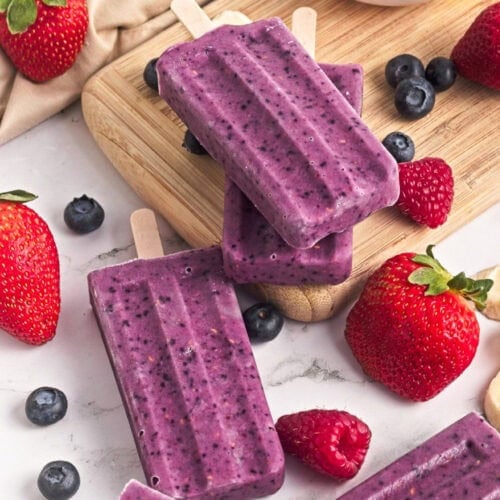 Triple berry smoothie popsicles stacked on a wooden board with fresh strawberries, blueberries, and raspberries.