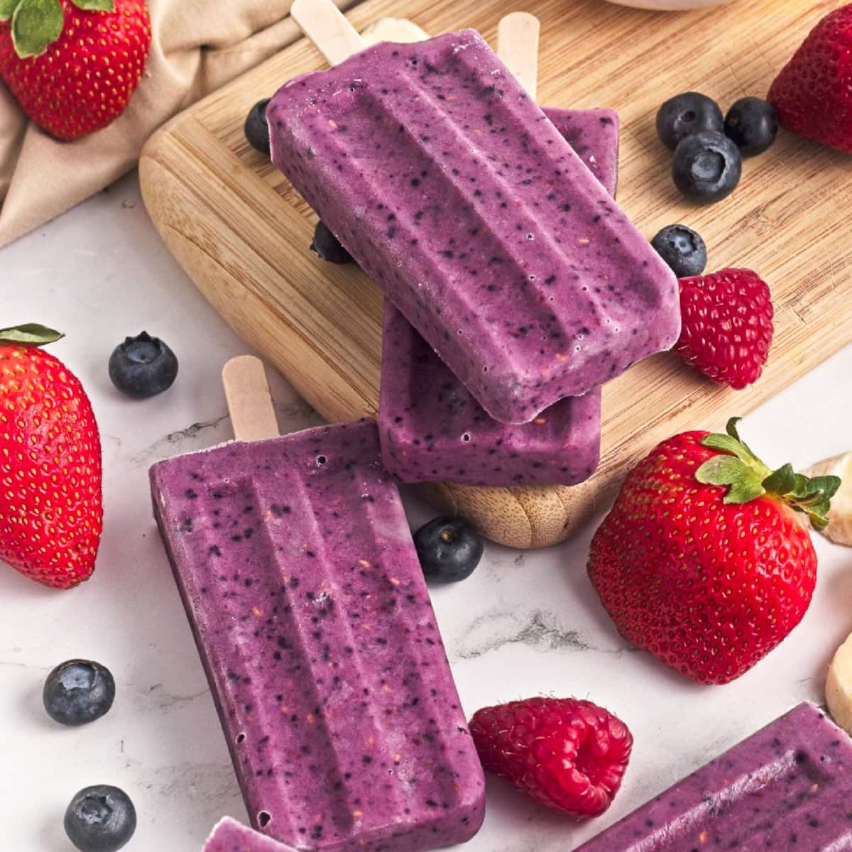 Triple berry smoothie popsicles stacked on a wooden board with fresh strawberries, blueberries, and raspberries.