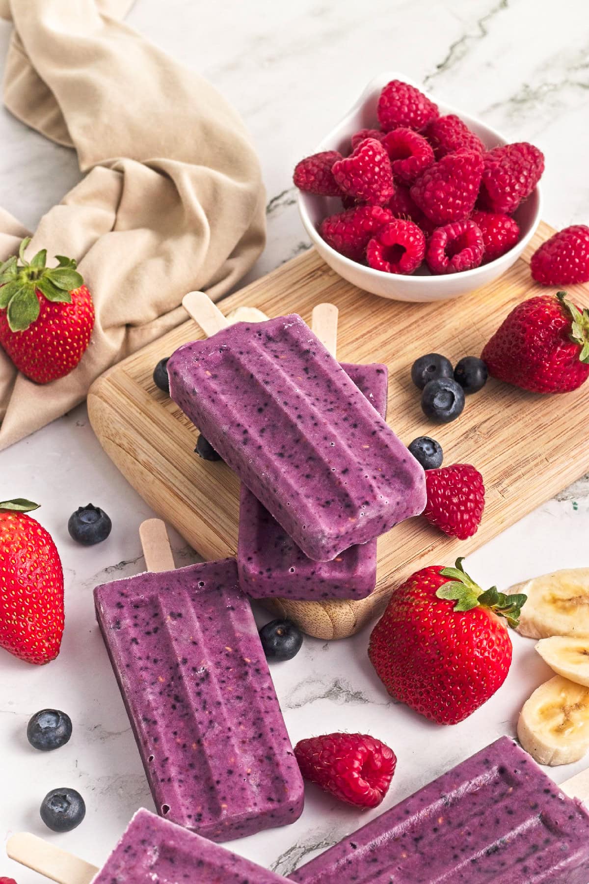 Close-up of triple berry smoothie popsicles stacked on a wooden board with fresh strawberries, raspberries, and blueberries.