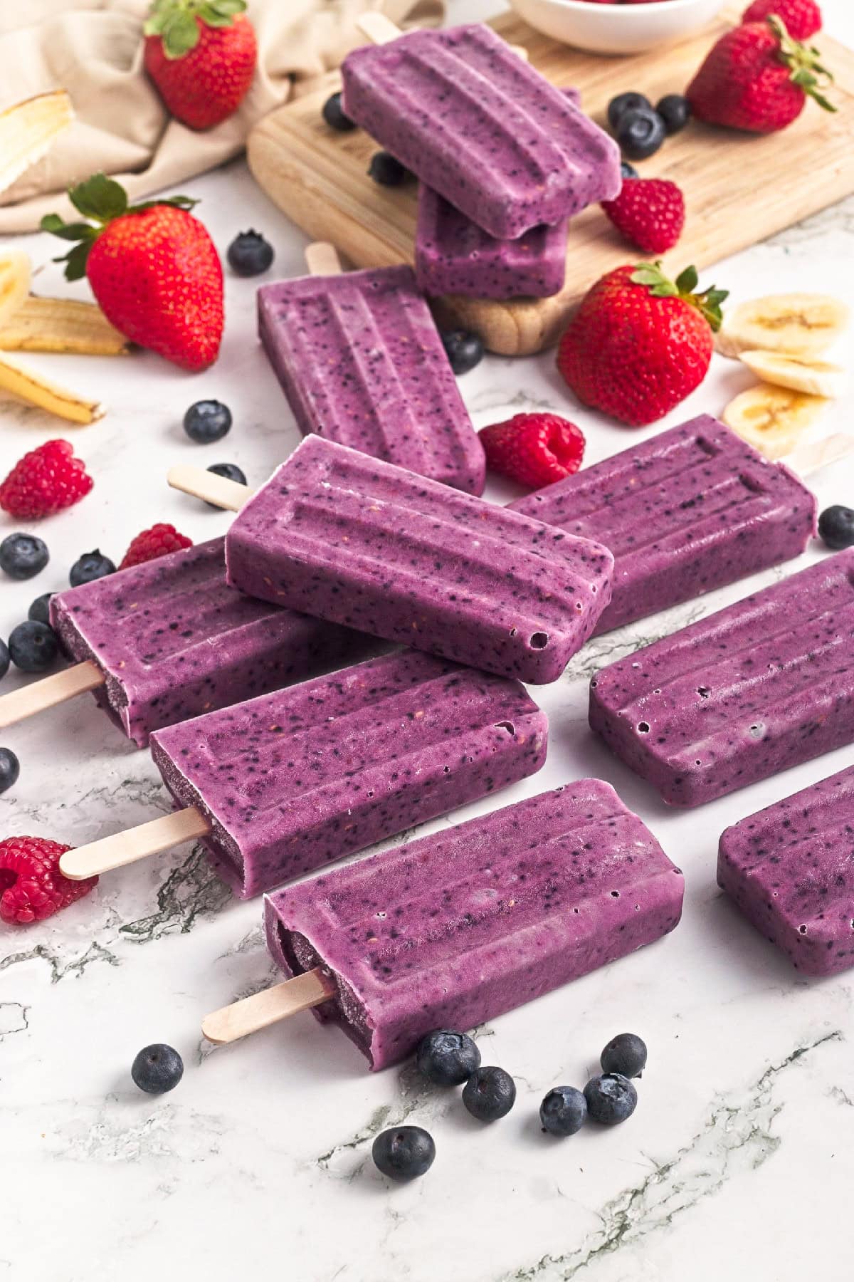 Several homemade triple berry smoothie popsicles served with fresh strawberries, blueberries, raspberries, and banana slices.