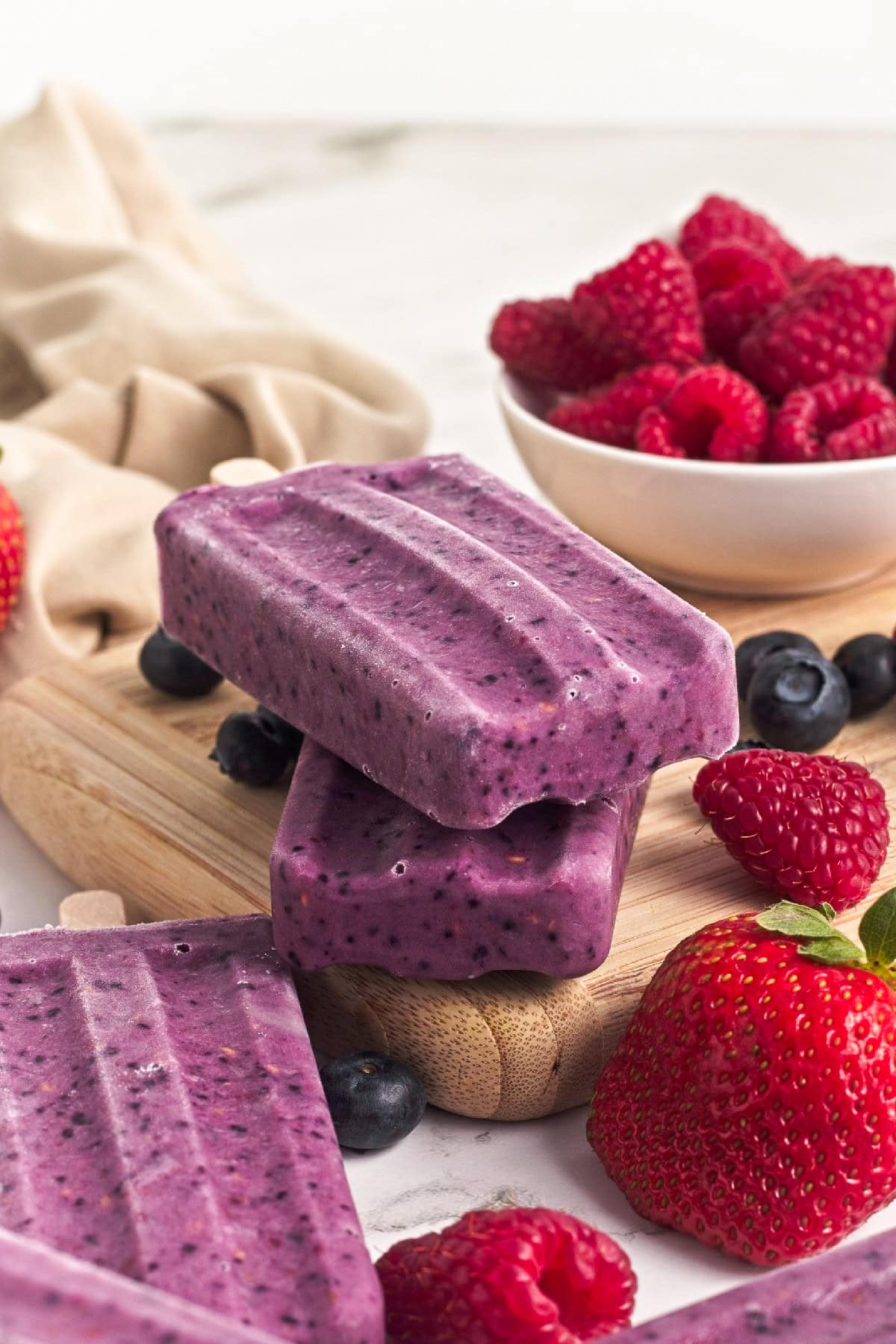 Stacked triple berry smoothie popsicles served with fresh strawberries, raspberries, and blueberries.