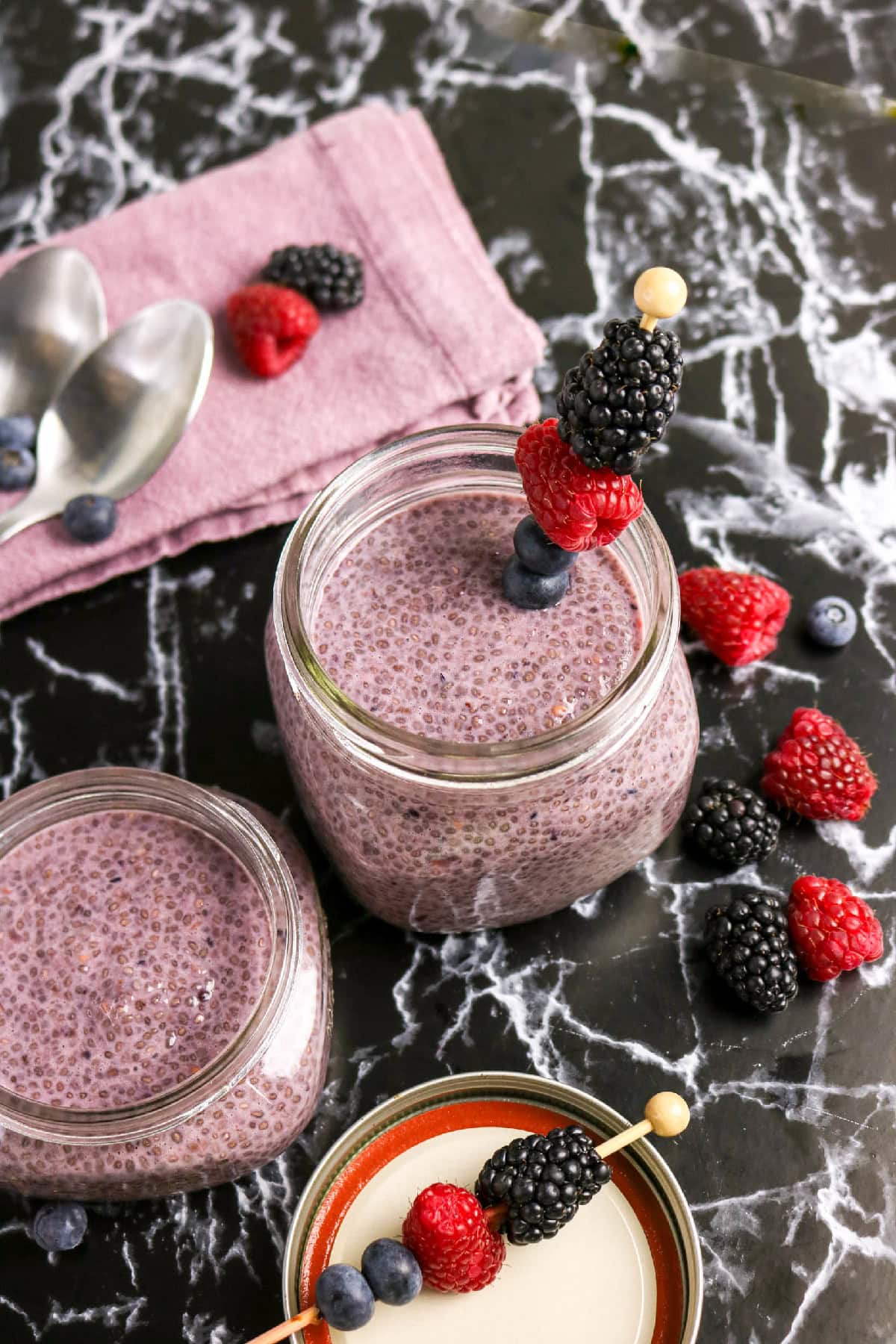 Berry chia pudding in glass jars garnished with skewered raspberries, blueberries, and blackberries.