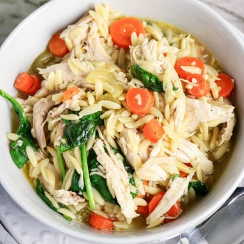 Chicken and orzo soup with shredded chicken, carrots, spinach, and celery in a white bowl.