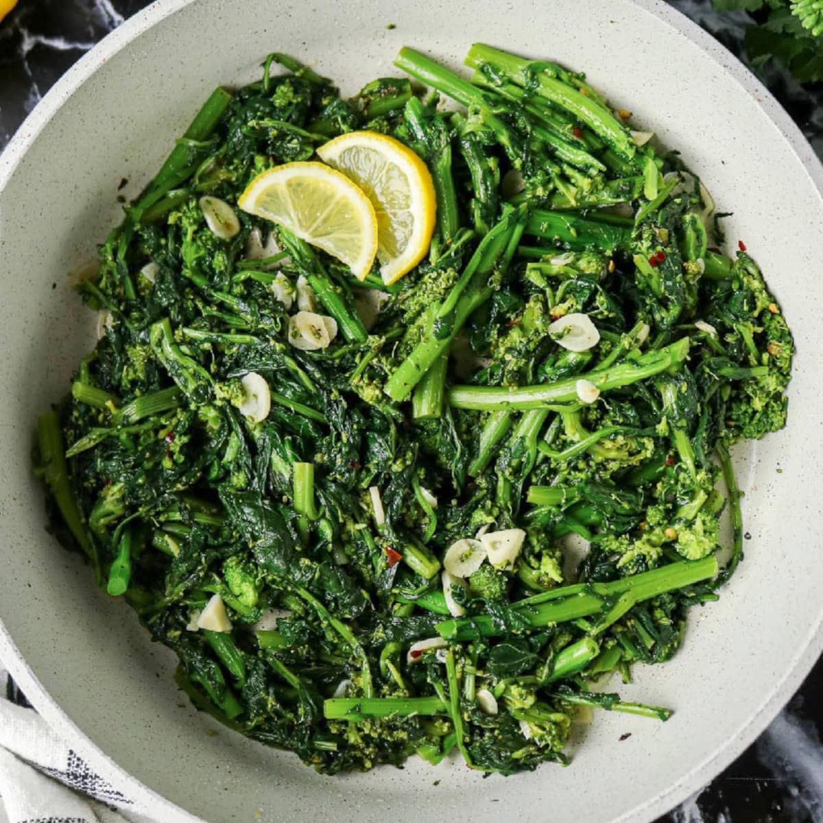 Sautéed rapini with sliced garlic, red pepper flakes, and fresh lemon wedges in a skillet.
