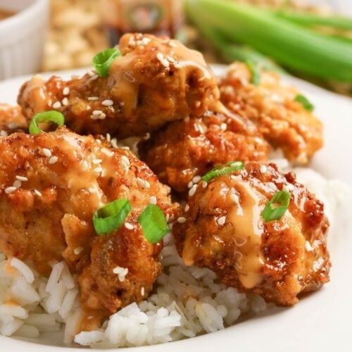 Close-up of crispy bang bang chicken drizzled with creamy sauce and topped with sesame seeds and green onions over rice.