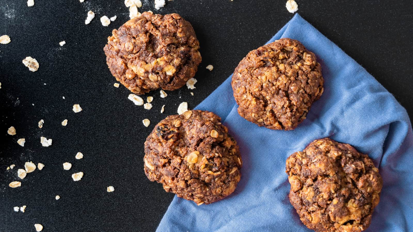 Four oatmeal cookies on a black surface with scattered oats and a blue cloth.