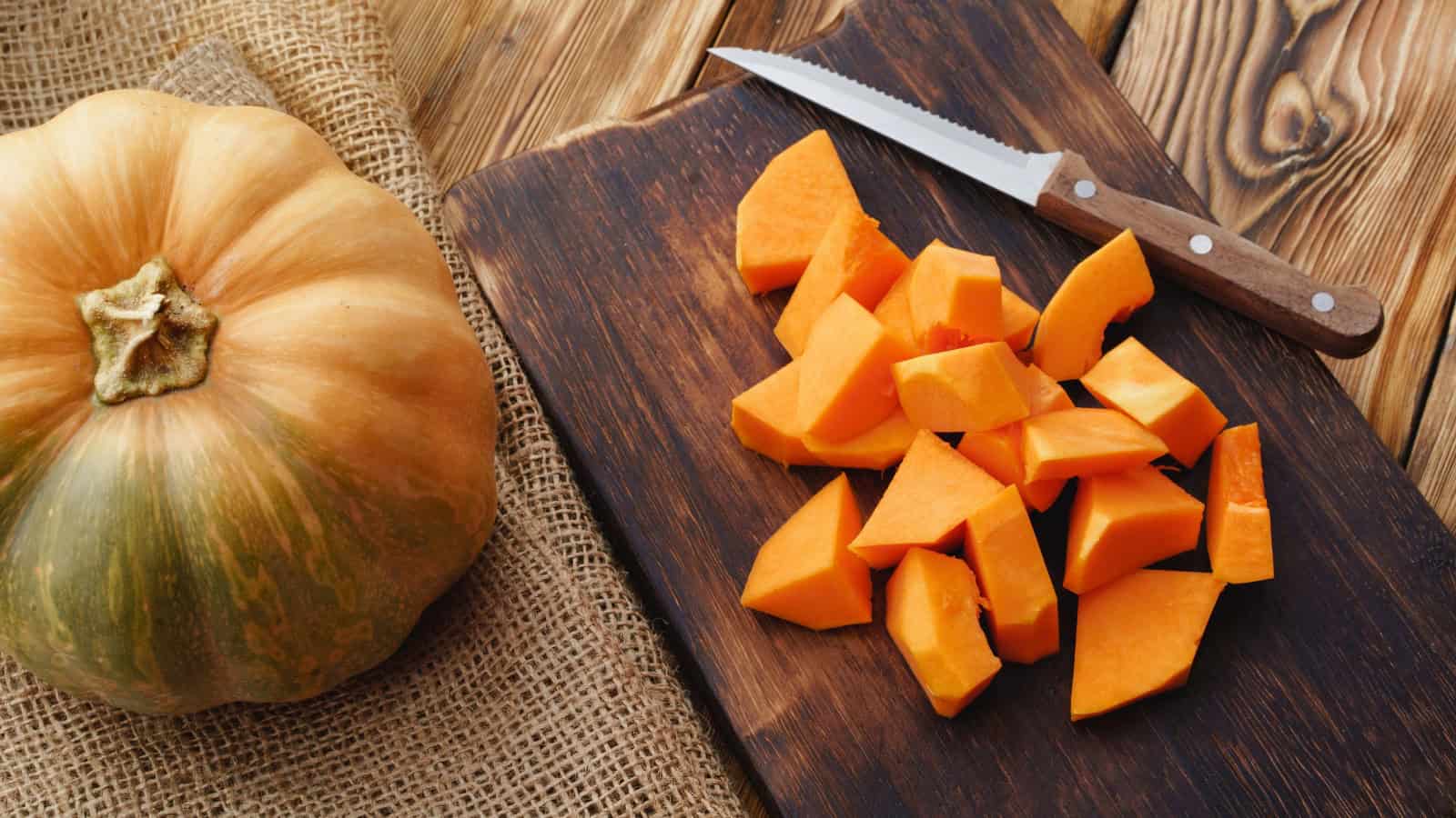 Whole pumpkin beside freshly cut pumpkin pieces on a wooden cutting board.
