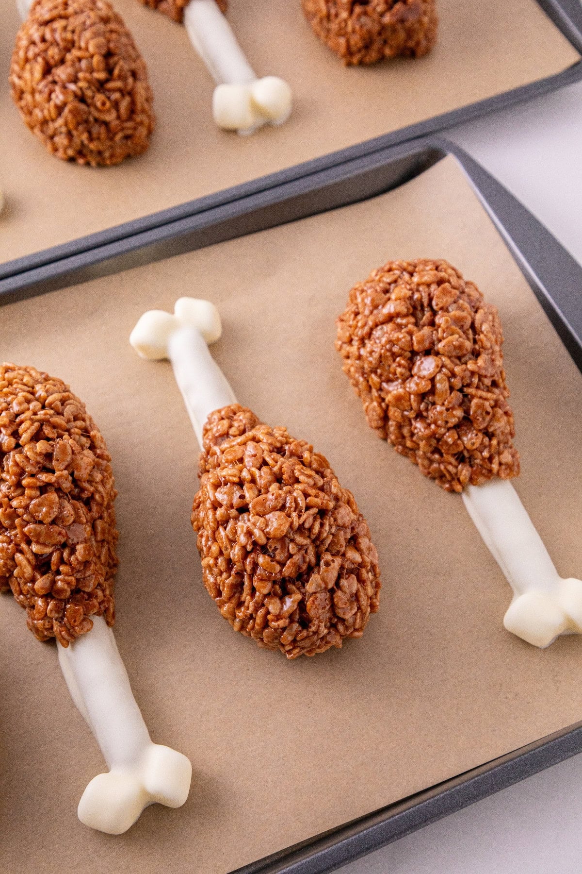 Rice Krispie turkey leg treats with white chocolate pretzel bones arranged on lined baking sheets.
