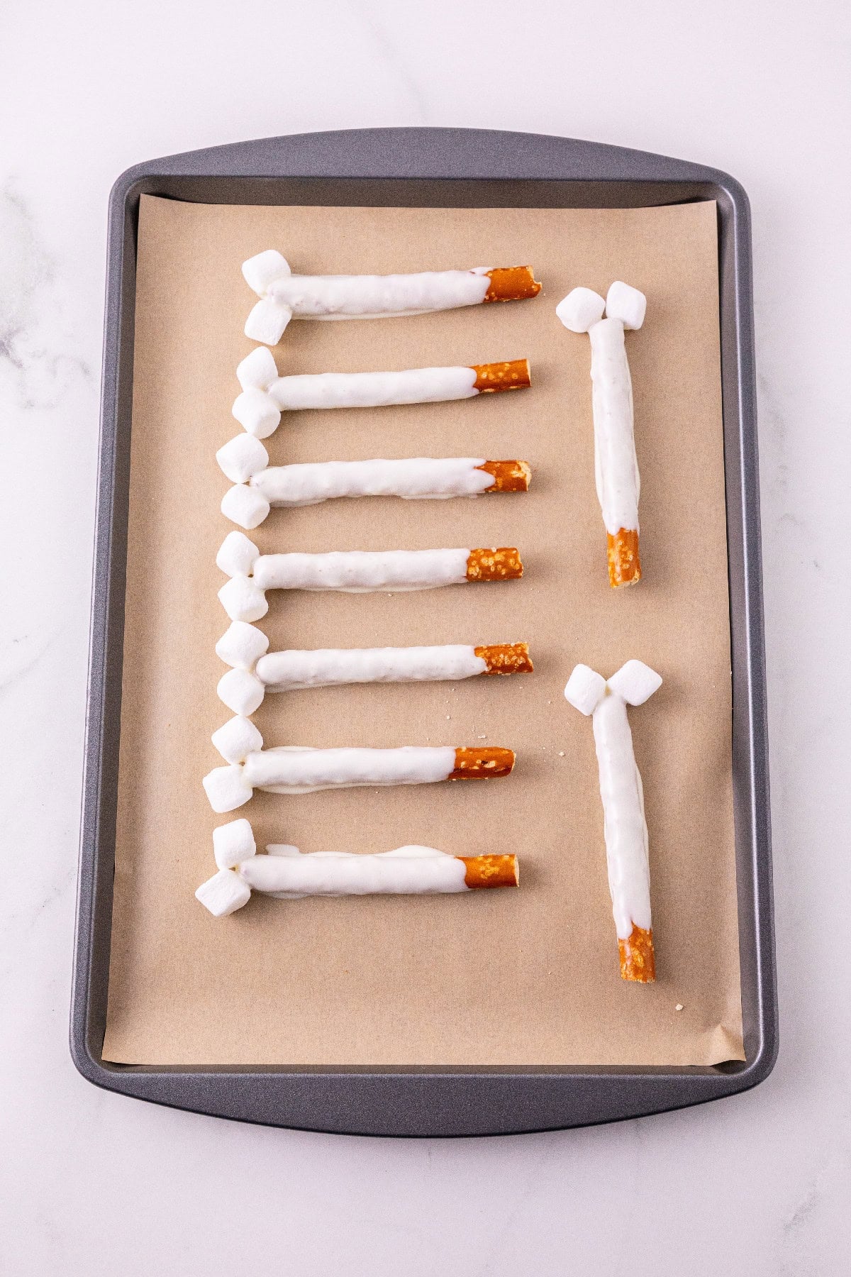 Dipped pretzel rods with mini marshmallows on a lined baking sheet.
