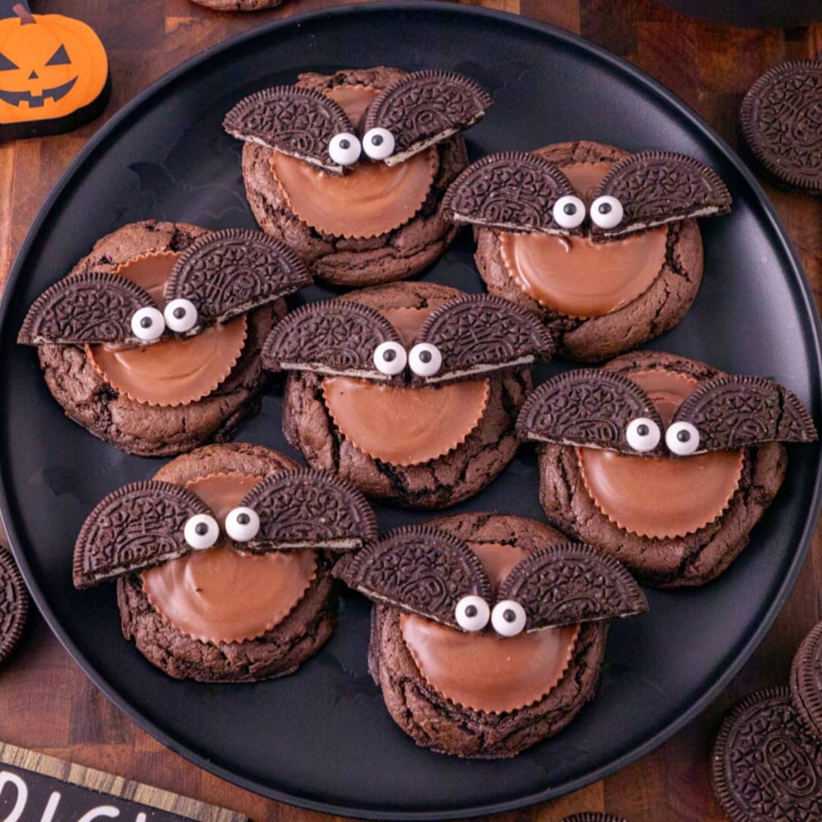 Oreo bat cookies topped with Reese’s peanut butter cups, Oreo wings, and candy eyes on a black plate.