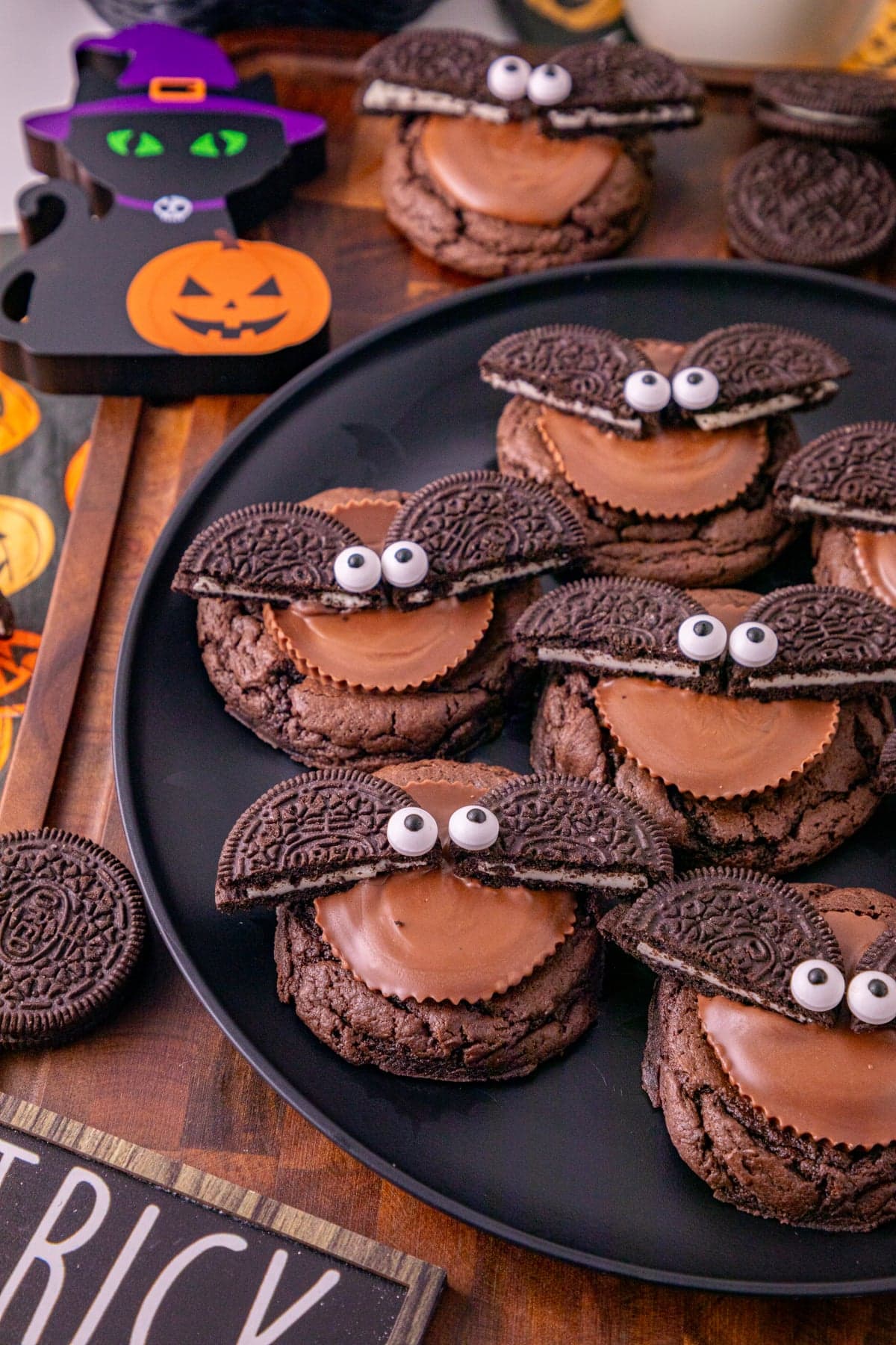 Oreo bat cookies with Reese’s peanut butter cups and candy eyes served on a black plate with Halloween decorations.