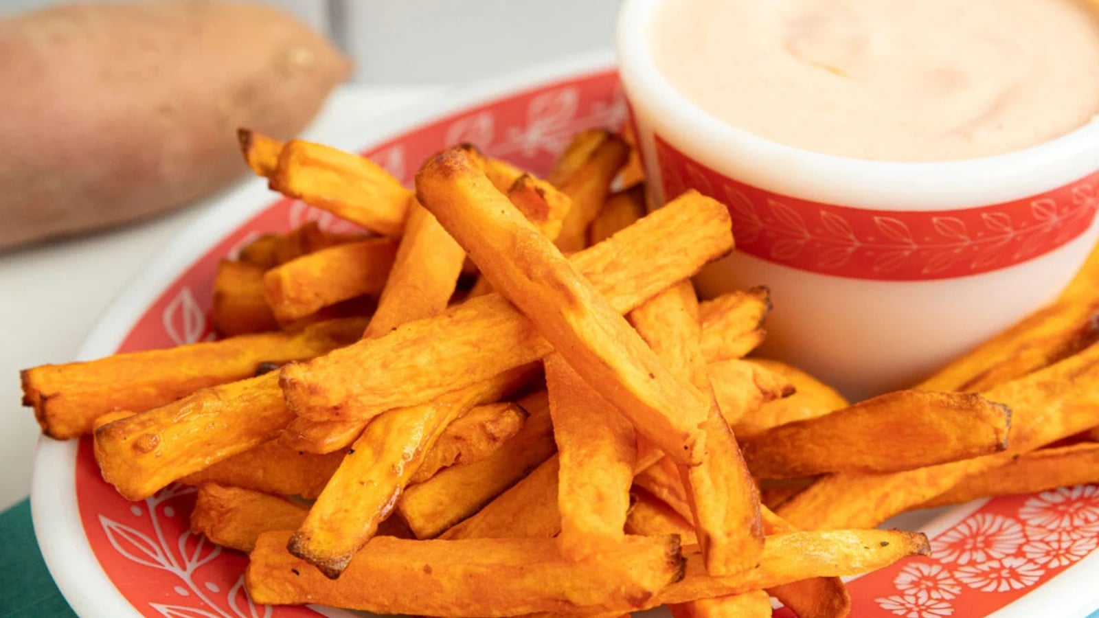 Crispy air-fryer sweet potato fries piled on a patterned plate next to a small bowl of dipping sauce.