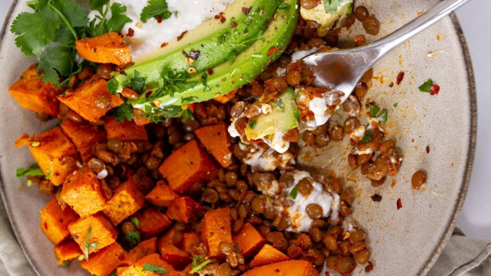 A savory bowl filled with roasted sweet potato cubes, brown lentils, cottage cheese, and sliced avocado, garnished with fresh cilantro and red pepper flakes.
