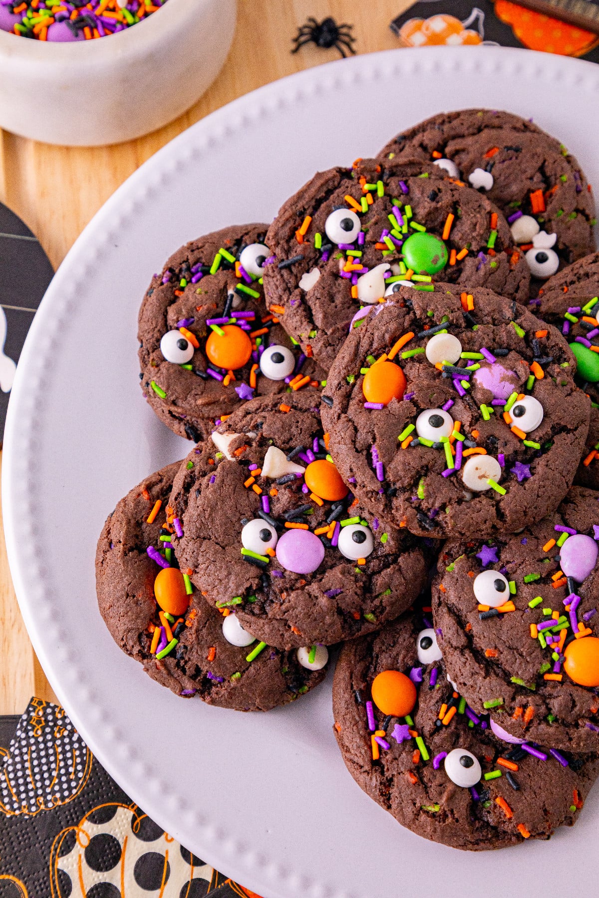 Close-up of chocolate Halloween cake mix cookies decorated with candy eyes, colorful sprinkles, and orange and green candies on a white plate.