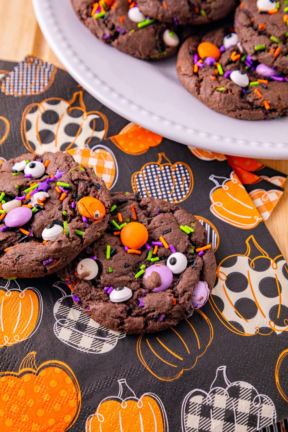 Two chocolate Halloween cake mix cookies topped with candy eyes, colorful sprinkles, and candies on a black pumpkin-patterned napkin.