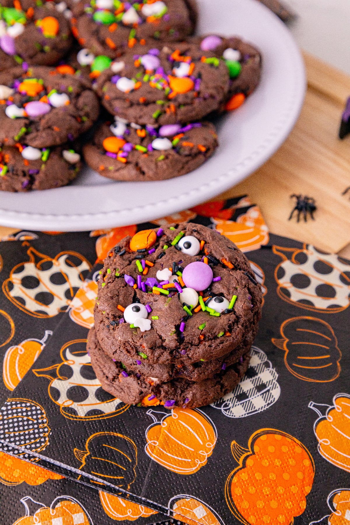 A chocolate Halloween cake mix cookie decorated with candy eyes and colorful sprinkles on a pumpkin-patterned napkin, with more cookies on a white plate in the background.