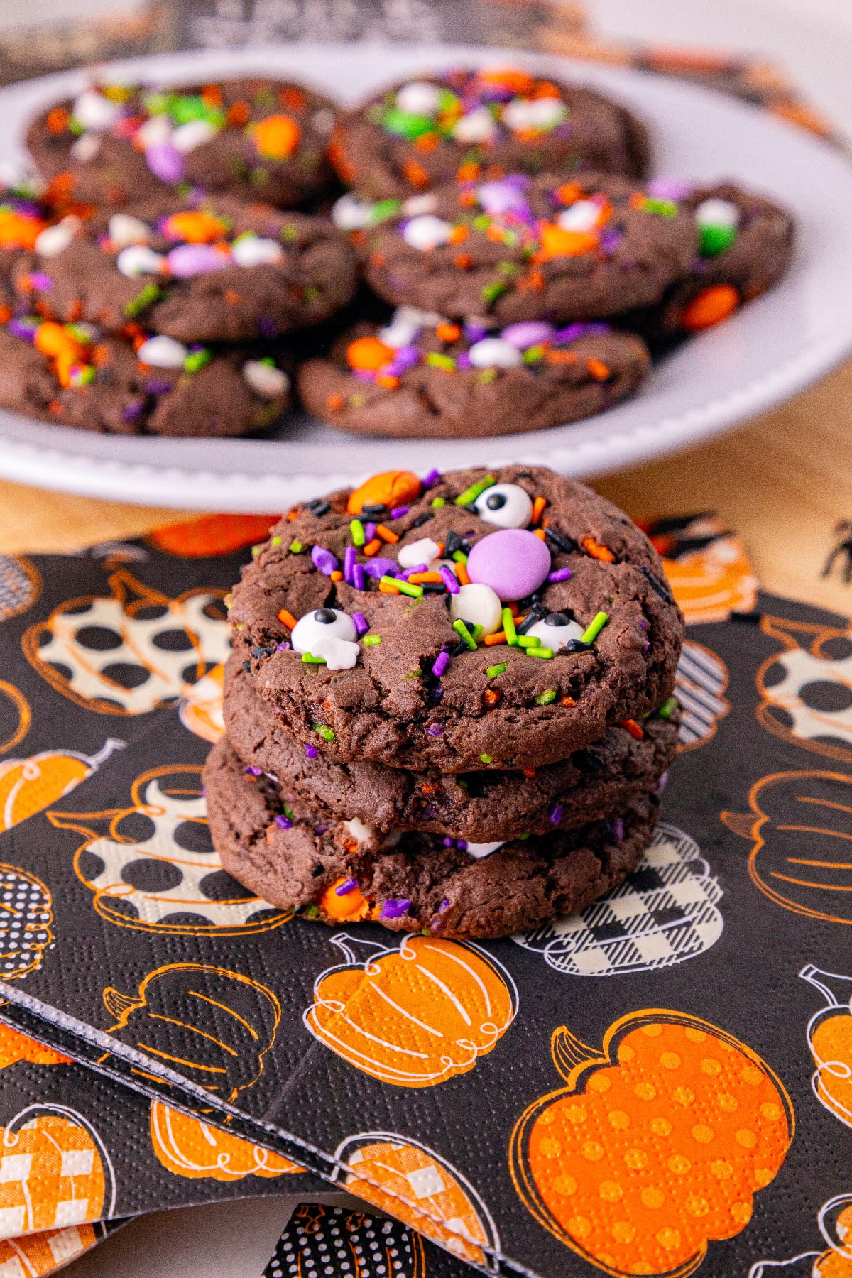 A stack of chocolate Halloween cake mix cookies with colorful sprinkles and candy eyes on a pumpkin-patterned napkin.
