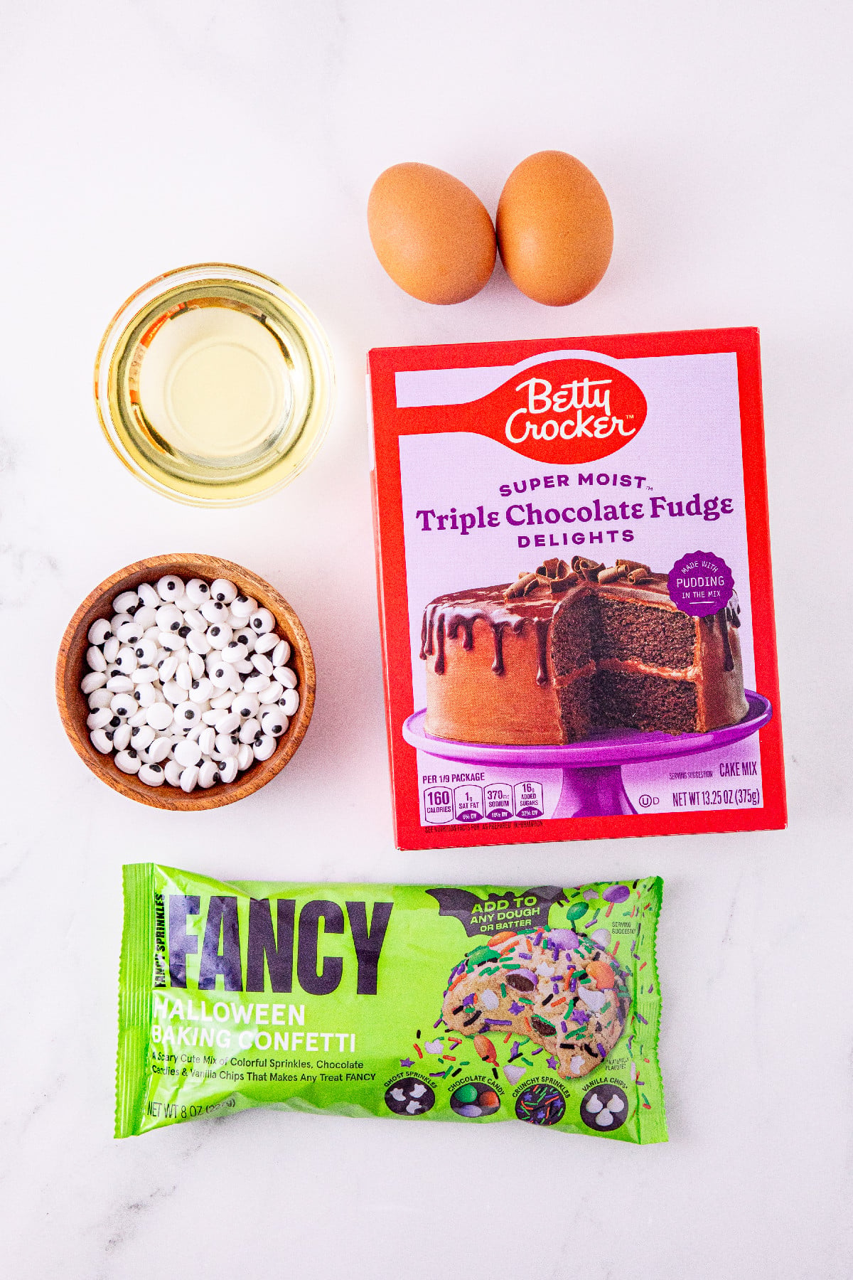 Ingredients for Halloween cake mix cookies, including chocolate cake mix, eggs, oil, candy eyes, and Halloween baking confetti.