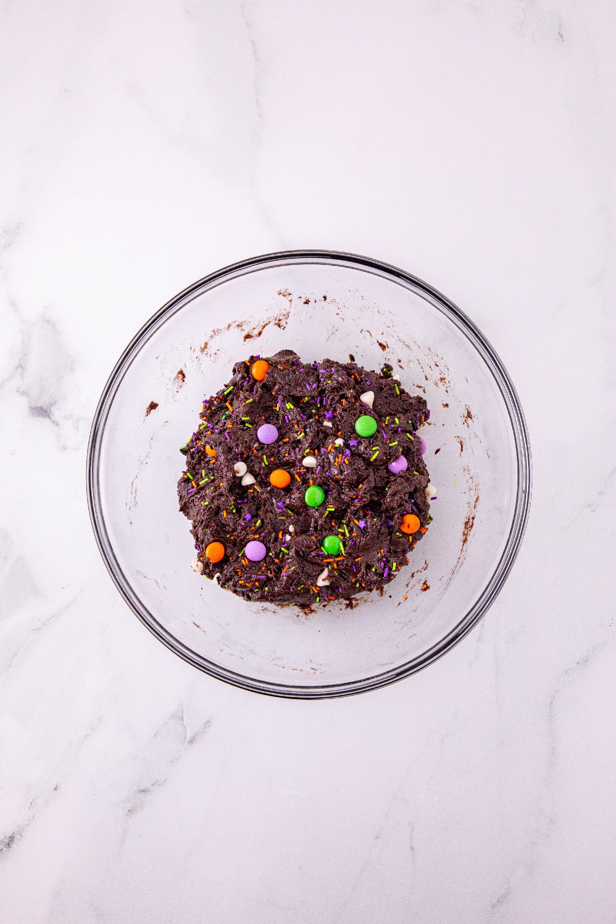 Chocolate cookie dough mixed with Halloween candy and sprinkles in a glass bowl.
