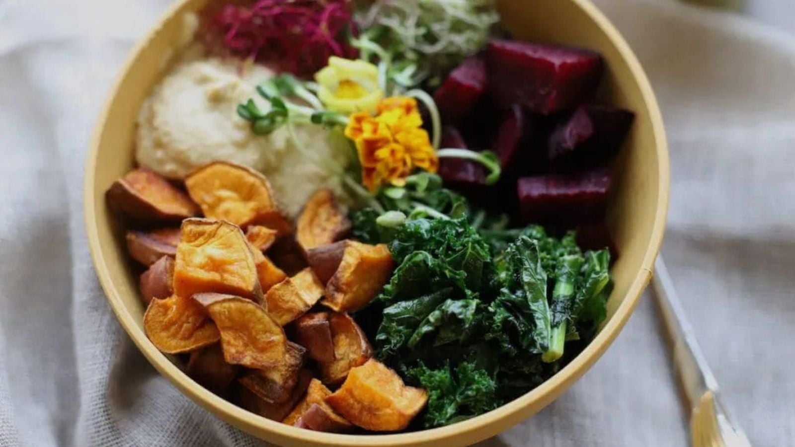 A colorful grain bowl with roasted sweet potato wedges, sautéed kale, diced beets, hummus, and sprouts, garnished with an edible flower.
