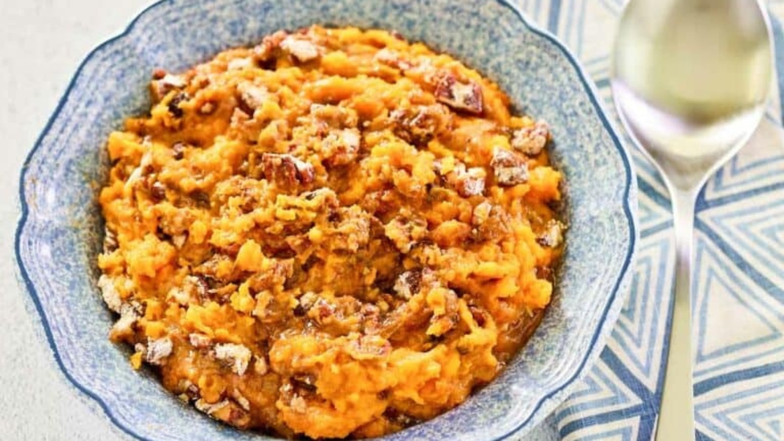 A blue serving bowl filled with mashed sweet potato casserole topped with a crunchy pecan crumble, served next to a metal spoon.