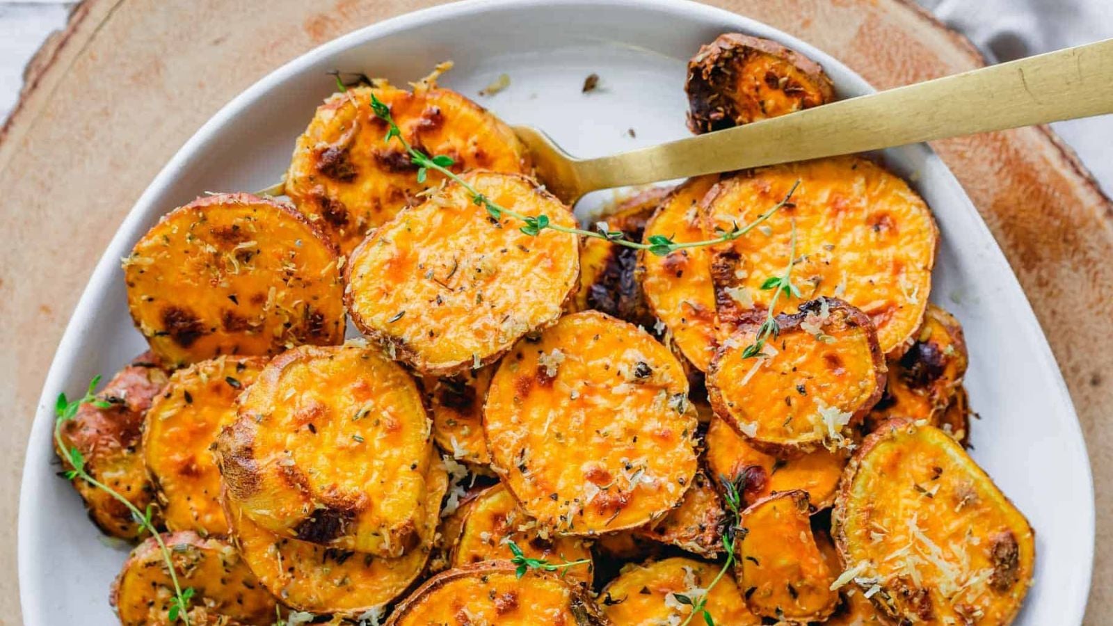 Roasted sweet potato rounds crusted with Parmesan cheese and fresh herbs, served in a shallow bowl with a gold serving spoon.
