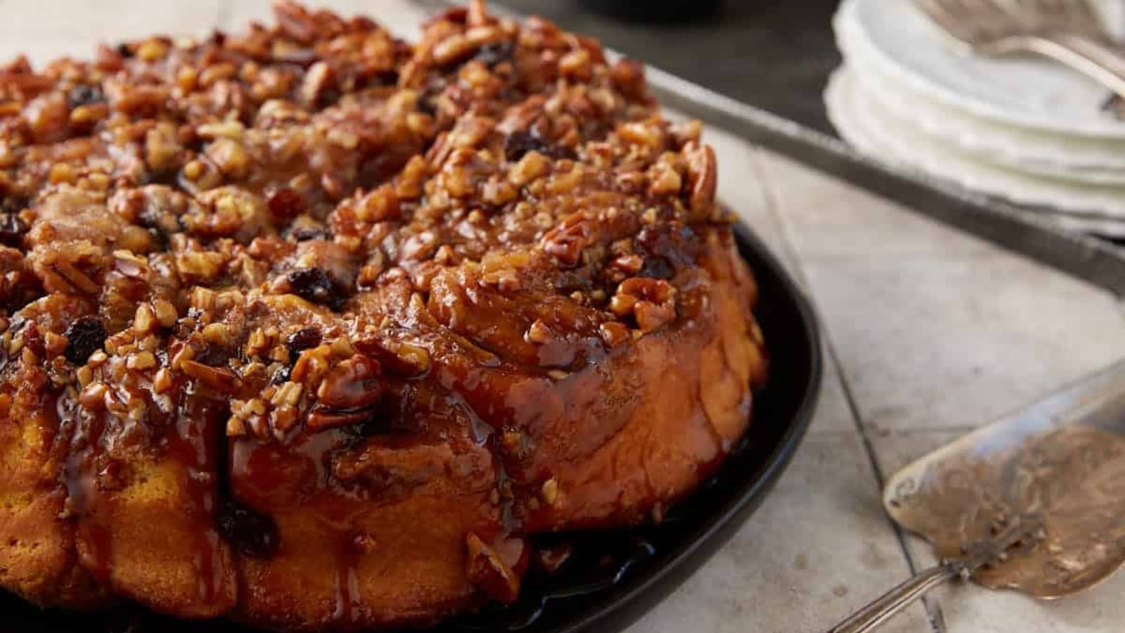 A round tray of sweet potato pecan sticky buns generously topped with chopped pecans, raisins, and a glossy caramel glaze.