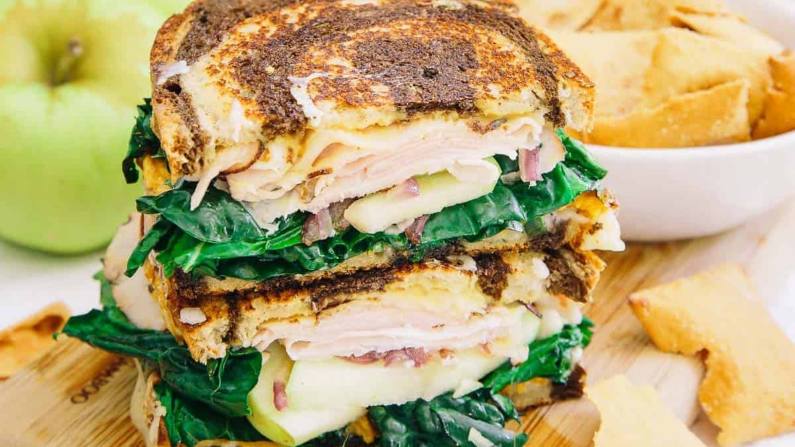 A stacked toasted sandwich on marble rye filled with sliced turkey, green apple slices, melted cheese, and leafy greens, served alongside a green apple and chips.