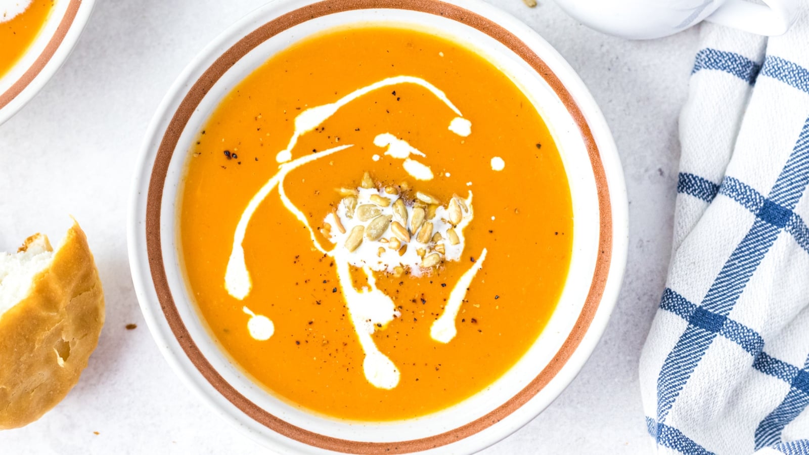 A bowl of creamy orange sweet potato soup garnished with a swirl of cream, cracked black pepper, and toasted sunflower seeds.