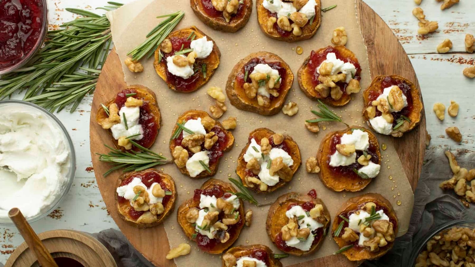 Roasted sweet potato rounds arranged on a wooden board, topped with cranberry sauce, goat cheese, toasted walnuts, and fresh rosemary sprigs.