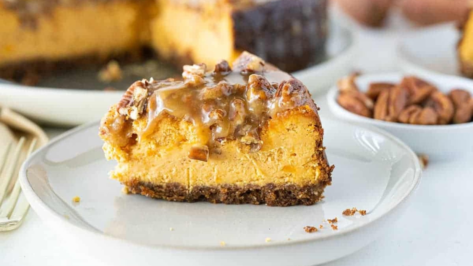 A slice of sweet potato cheesecake on a graham cracker crust, topped with chopped pecans and caramel drizzle.