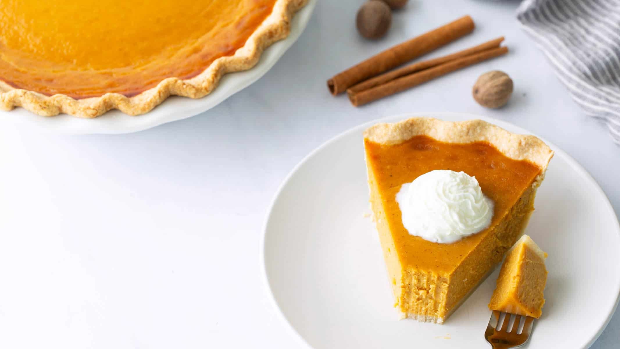 A slice of sweet potato pie topped with whipped cream served on a plate next to whole spices and a full pie.