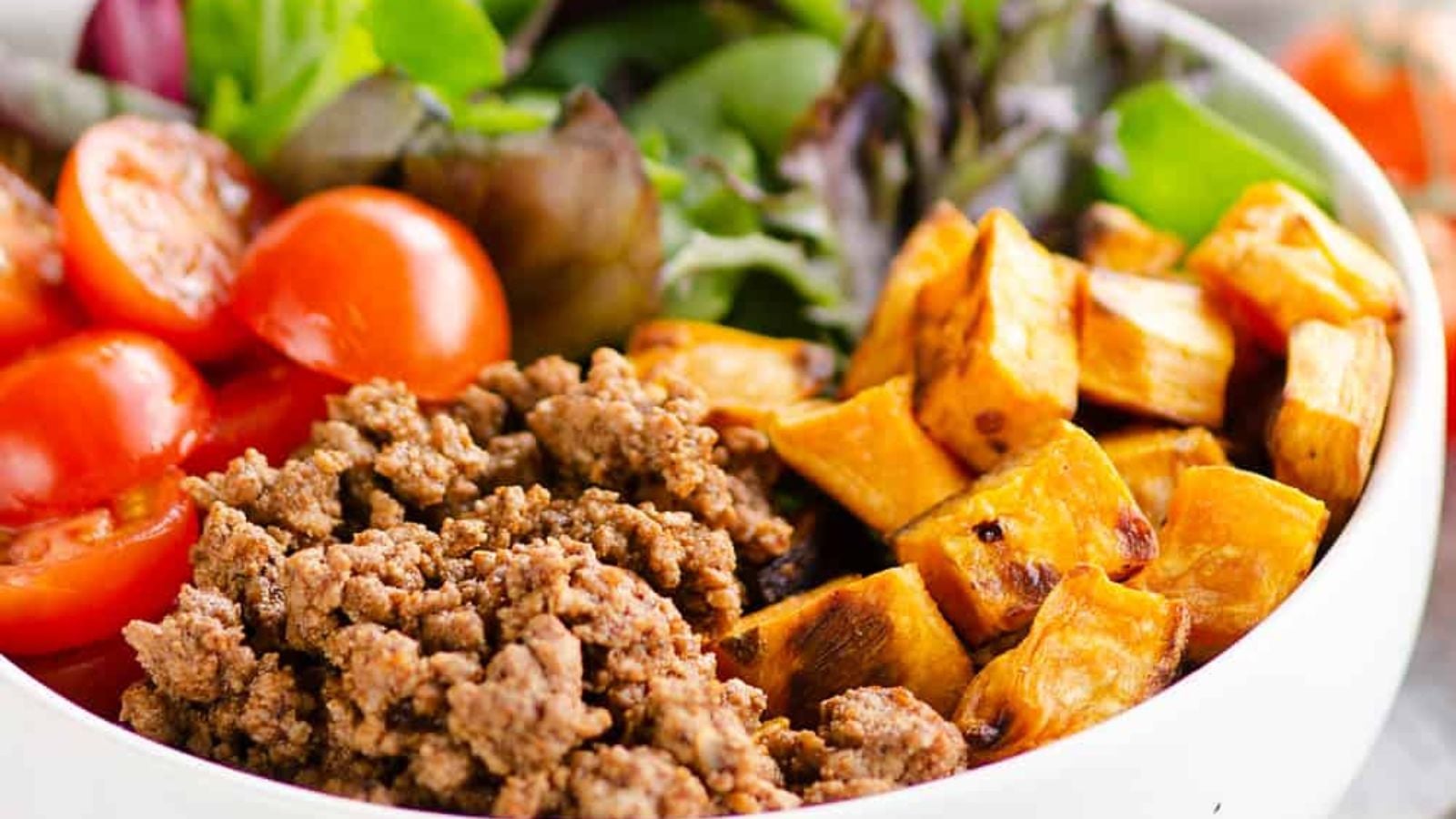 A taco bowl loaded with seasoned ground meat, roasted sweet potato cubes, cherry tomato halves, and mixed greens in a white bowl.