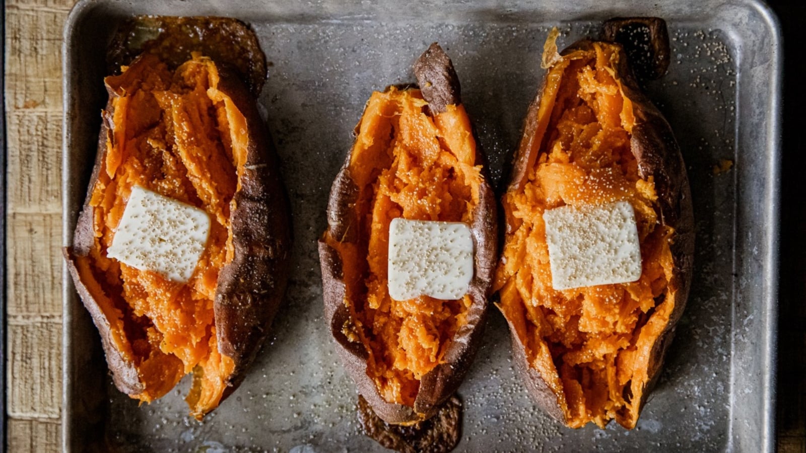 Three baked sweet potatoes split open on a baking sheet, each topped with a pat of butter and a sprinkle of spices.
